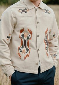 Beige gestreept shirt met geometrische borduursels in roest, zwart en bruine tinten. Knopsluiting met een klassieke kraag en manchetten.