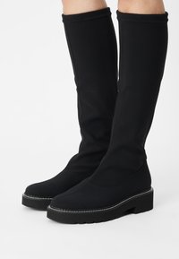 Botas negras hasta la rodilla hechas de un tejido elástico con tacón elevado, que presentan suelas planas y costuras en contraste a lo largo de los bordes.