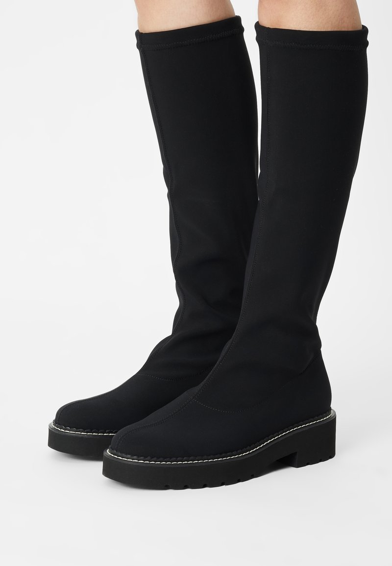 Botas negras hasta la rodilla hechas de un tejido elástico con tacón elevado, que presentan suelas planas y costuras en contraste a lo largo de los bordes.