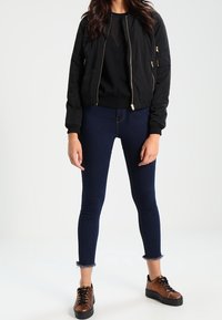 Giacca bomber nera con cerniere dorate, abbinata a jeans skinny blu scuro con orli sfrangiati e sneakers marroni con lacci.