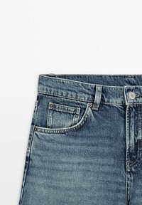 Section de la taille d'un jean en denim bleu avec bouton avant, passants de ceinture et deux poches avant, présenté sur fond blanc.