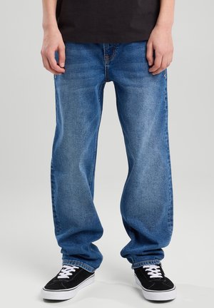 Blue denim jeans with a loose fit, featuring five pockets and subtle fading. Worn with black sneakers with white laces and a classic design.