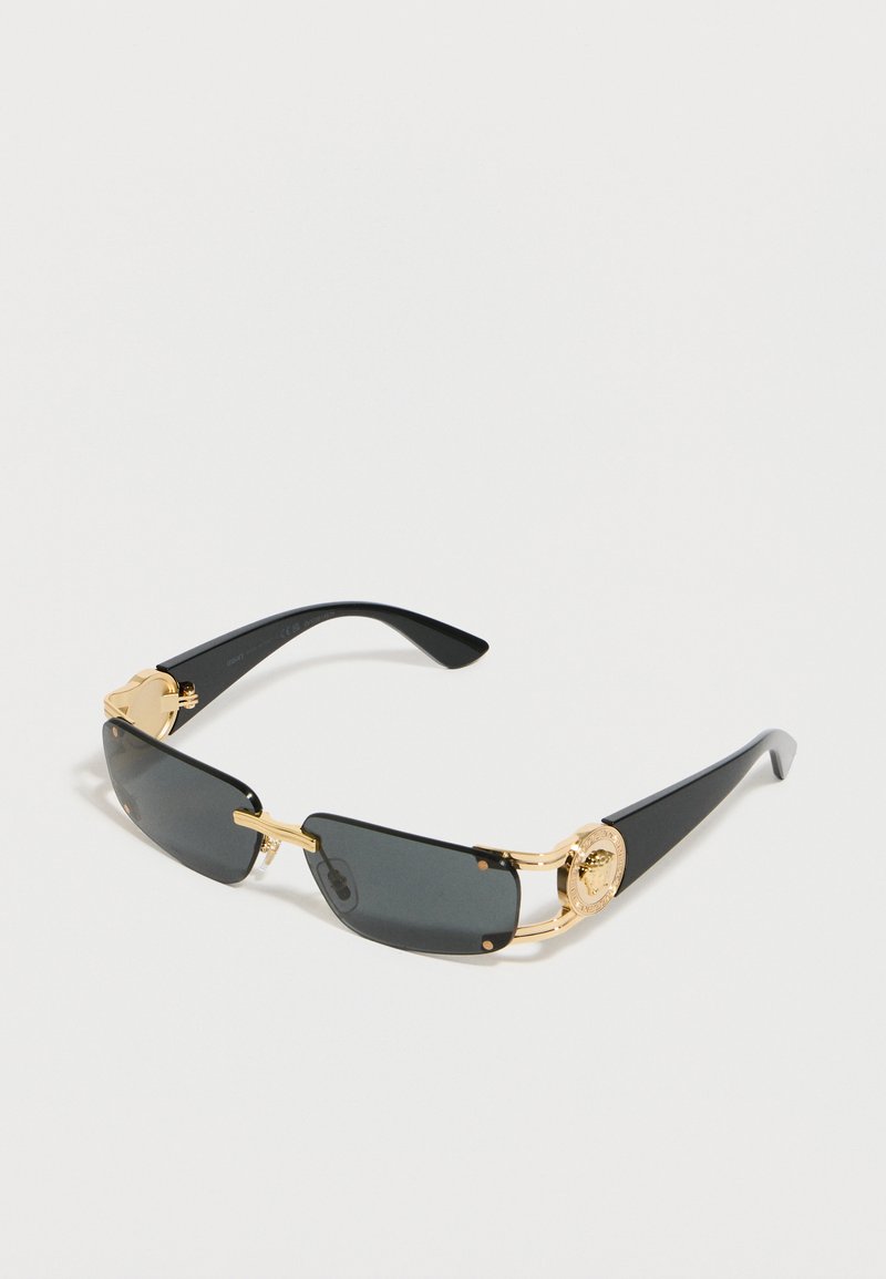 Ορθογώνια μαύρα γυαλιά ηλίου με χρυσές λεπτομέρειες και διακοσμητικά χρυσά μετάλλια στα μαύρα σκέλη, εμφανίζονται μπροστά σε φωτεινό φόντο.