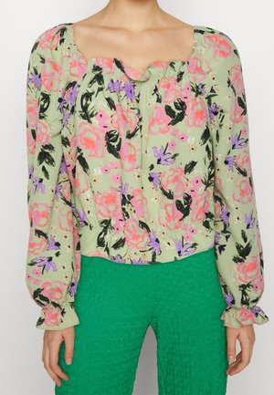 Mujer con blusa verde con flores rosas y moradas y mangas largas abullonadas, combinada con pantalones texturizados verde brillante.