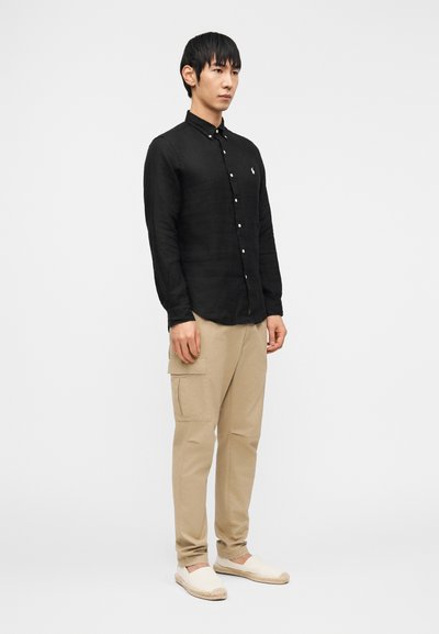 Bărbat în picioare, purtând o cămașă neagră cu nasturi, pantaloni cargo bej și pantofi slip-on albi, pe un fundal alb simplu.