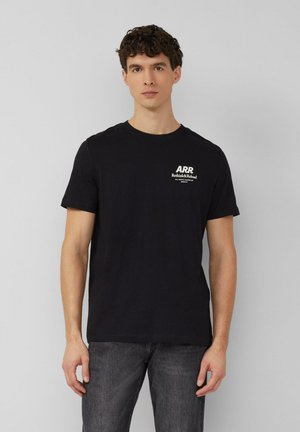 Młody mężczyzna w czarnej koszulce z krótkim rękawem z białym napisem "ARR Rethink&Reload" na piersi, w ciemnoszarych dżinsach, stoi na tle szarej ściany.