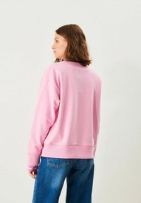 Kvinna med axellångt brunt hår som bär en rosa sweatshirt och blå jeans, sedd i profil mot en enfärgad bakgrund.
