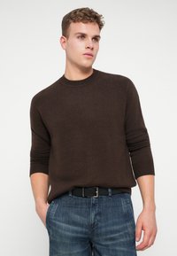 Ung man med kort, lockigt hår som bär en mörkbrun långärmad tröja och blå jeans, står med en hand i fickan och tittar åt vänster.