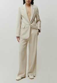Beige karierter Blazer mit tiefem V-Ausschnitt, Einzelknopfverschluss und weit geschnittenen Hosen. Der Stoff ist glatt, leicht und maßgeschneidert.