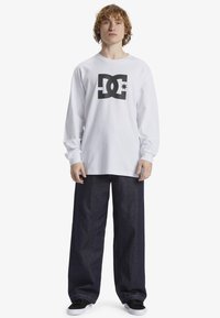 Long-sleeve white t-shirt with a black DC logo on the chest, paired with loose dark denim jeans and black sneakers.
