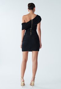 Zwarte off-shoulder jurk met geraffineerde stof, getailleerde pasvorm en zichtbare gouden rits aan de achterkant. Draagt met schouderbandjes en hoge hakken.