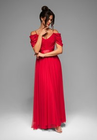 Robe maxi rouge à épaules dénudées avec un corsage froncé et une jupe fluide. Tissu transparent avec un aspect texturé. Talons argentés à lanières visibles.