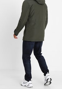 Olijfgroene gewatteerde jas met een gladde textuur, gecombineerd met donkerblauwe spijkerbroek en witte sportschoenen, gezien van achteren.