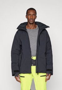 Mies, joka käyttää mustaa vetoketjullista takkia harmaan neuleen päällä ja kirkkaan keltaisia housuja, joissa on mustia vetoketjuyksityiskohtia, seisoo valkoista taustaa vasten.