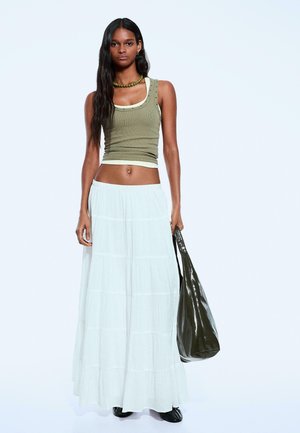 Olivgrünes Tanktop mit dekorativen Nieten, kombiniert mit einem gestuften weißen Maxikleid und einer dunkelgrünen Tragetasche. Schwarze Stiefeletten runden den Look ab.