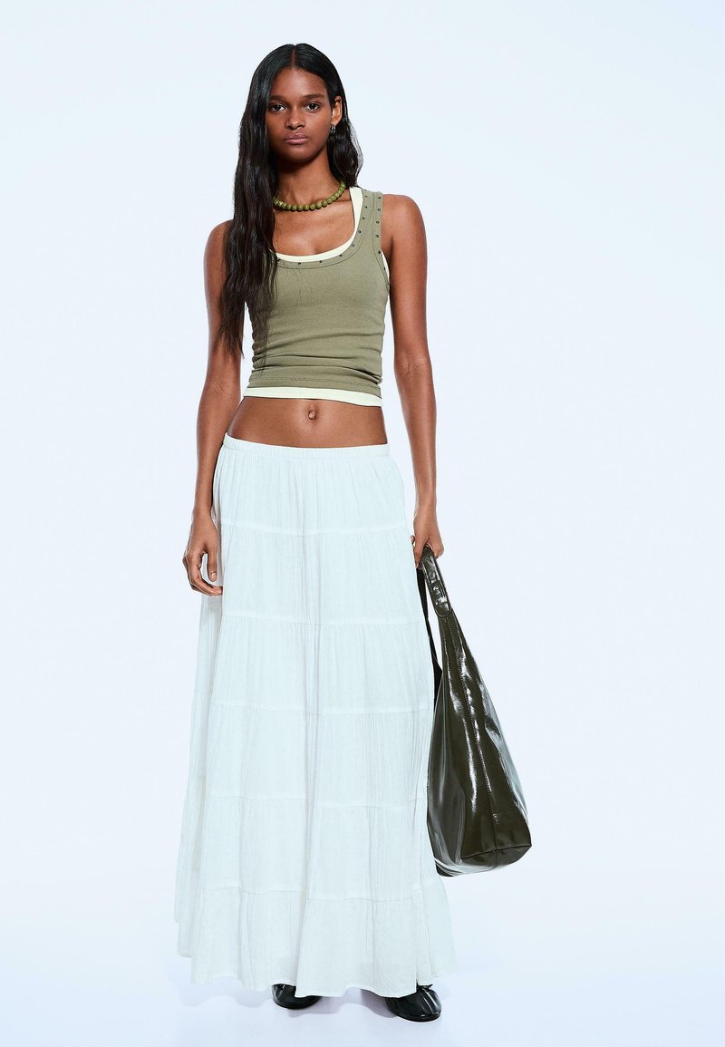 Olivgrünes Tanktop mit dekorativen Nieten, kombiniert mit einem gestuften weißen Maxikleid und einer dunkelgrünen Tragetasche. Schwarze Stiefeletten runden den Look ab.
