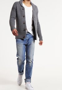 Osoba kráčející, oblečená v šedém pleteném kardiganu na knoflíky, bílém tričku, modrých džínách s ohrnutými lemy a bílých teniskách.