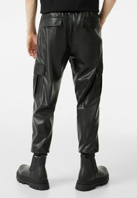 Svarta byxor i konstläder med elastisk midja, två sidofickor och ankellånga fållar. Kombineras med kraftiga svarta kängor.