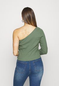 Green, long-sleeve top with one shoulder exposed. Made of ribbed fabric, features a fitted design, worn with blue jeans.