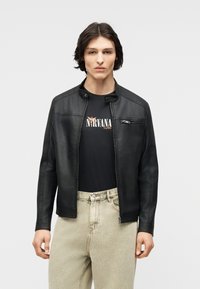 Young person wearing a black leather jacket over a black Nirvana t-shirt and light beige jeans, standing against a plain background.