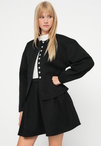 Jachetă neagră structurată cu nasturi mari albi, asortată cu o fustă neagră corespunzătoare. Materialul pare neted cu o ușoară textură.
