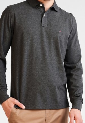 Homme portant un polo à manches longues gris foncé avec deux boutons blancs et un petit logo sur la poitrine, associé à un pantalon beige.