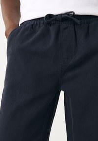 Vyras, dėvintis tamsiai mėlynas raišteliu suveržtas kelnes su elastine liemene ir baltą marškinių, rankos kišenėse, vaizduojamas nuo liemens iki vidurio šlaunų.