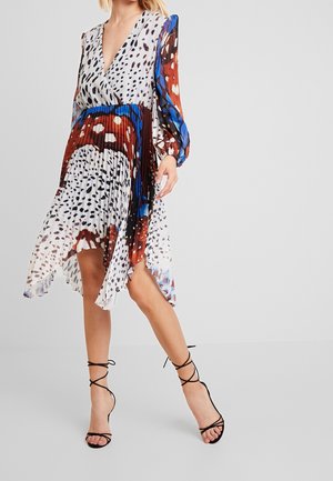 Woman wearing a pleated, multicolored animal print dress with long sleeves and black lace-up high-heeled sandals, standing on a gray floor.