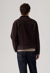 Bruine corduroy jas met een klassieke snit, voorzien van een kraag, knoopsluitingen bij de mouwen en zakken, aangevuld met een gebrokenwitte trui eronder.