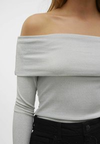 Sidabrinė off-the-shoulder palaidinė su ilgomis rankovėmis, turinti blizgų paviršių ir prigludusį siluetą, derinama su aukštu liemeniu juodomis džinsomis.
