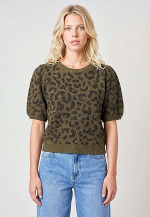 Blond vrouw met een olijfgroen kortemouwen trui met zwarte luipaardprint en lichtblauwe hoge taille jeans, staand tegen een effen achtergrond.