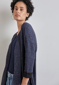 Marinblå öppen cardigan i texturerad tyg med svarta manschetter, bärs över en marinblå topp, med lös passform och ärmar av medellängd.