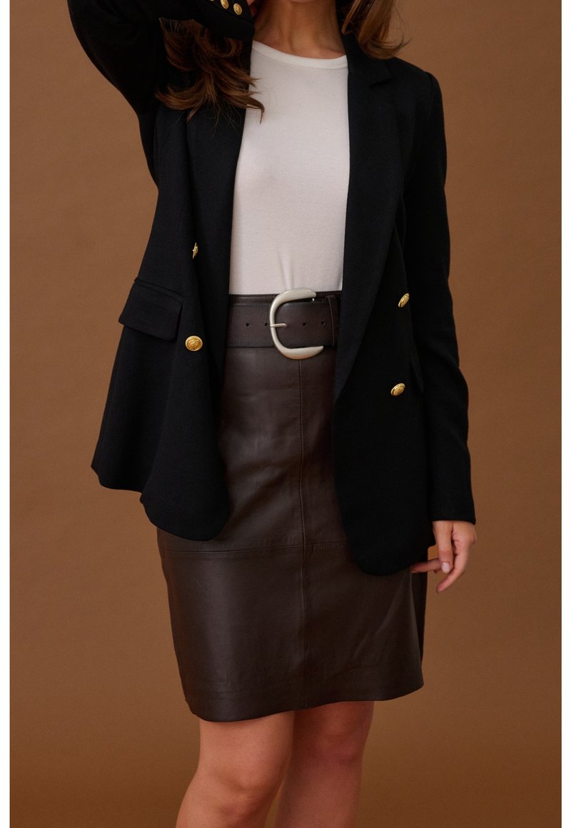 Femme portant un blazer noir avec des boutons dorés, un haut blanc, une jupe en cuir marron foncé et une large ceinture noire avec une boucle argentée.