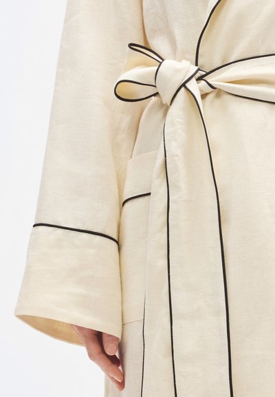 Gaišas bēša lina halāts ar melnām līnijām. Tam ir saite jostasvietā un sānu kabata, izceļot vaļīgu piegriezumu un gludu tekstūru.