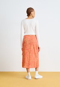 Rød-oransje skjørt med blomsterdesign, lett stoff, midtlengde. Kombinert med en hvit, ribbet langermet topp og hvite joggesko.