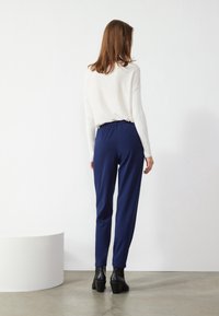 Marina smala byxor med elastisk midja, i kombination med en vit långärmad topp, bärs med svarta klackstövlar. Släta texturer visas.