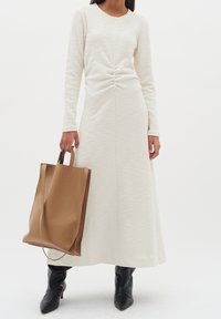 Vit långärmad klänning i texturerat tyg, med en rynkad detalj vid midjan. Modellen håller en brun läderhandväska och bär svarta ankelstövlar.