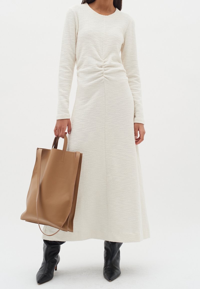 Vit långärmad klänning i texturerat tyg, med en rynkad detalj vid midjan. Modellen håller en brun läderhandväska och bär svarta ankelstövlar.