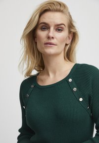 Maglione a coste verde scuro con scollatura arrotondata e bottoni decorativi in argento lungo la cucitura della spalla. Texture morbida e design aderente.
