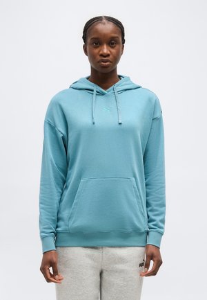 Γυναίκα με πλεξούδες, που φοράει ανοιχτό μπλε φούτερ με κουκούλα και ανοιχτό γκρι παντελόνι φόρμας, στέκεται μπροστά σε έναν απλό φόντο.