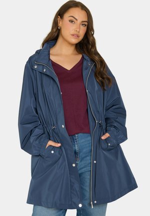 Donna con capelli lunghi e castani che indossa un impermeabile blu con cappuccio sopra una maglietta bordeaux a scollo a V e jeans blu, con le mani nelle tasche del cappotto.