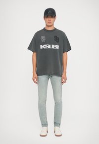 Gray oversized T-shirt with graphic print and logo, paired with light blue distressed jeans and white sneakers. Black cap completes the look.
