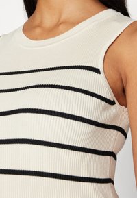 Ribbad linne i krämfärg med svarta horisontella ränder, har en rund halsringning och är ärmlös, vilket framhäver den texturerade tyget.