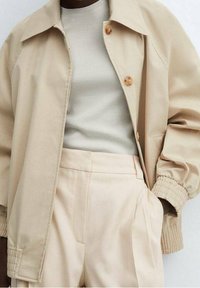 Beige jacket with a collar, large buttons, and elastic cuffs, layered over a light ribbed top and high-waisted pleated trousers.