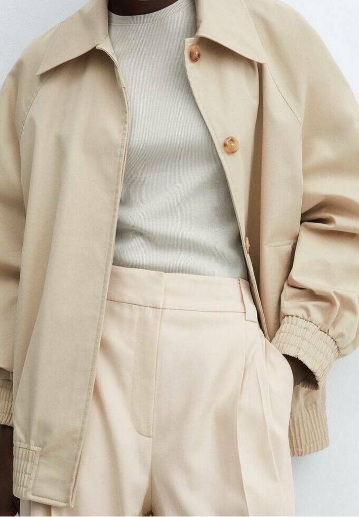Beige jacket with a collar, large buttons, and elastic cuffs, layered over a light ribbed top and high-waisted pleated trousers.