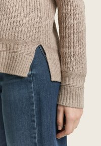 Beige knitted sweater with ribbed cuffs and hem, featuring a side slit. Worn over dark blue denim jeans. Texture appears chunky and cozy.