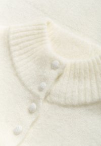 Közeli kép egy törtfehér kötött pulcsi gallérjáról, melyen bordázott rész és négy szövettel bevont gomb található a vállvarrás mentén.
