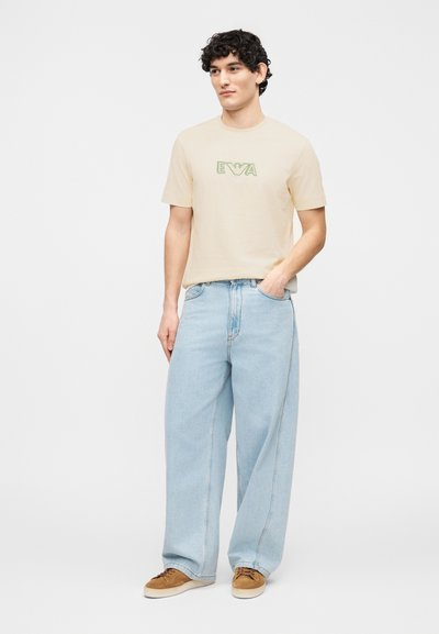 Νέος με σγουρά μαλλιά, ντυμένος με μπεζ κοντομάνικο μπλουζάκι με το λογότυπο "EVA", ανοιχτό μπλε τζιν με φαρδιά πόδια και καφέ αθλητικά παπούτσια, στέκεται με το ένα χέρι στην τσέπη.