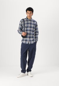 Καρό πουκάμισο σε μπλε, λευκό και γκρι, συνδυασμένο με cargo παντελόνι σε σκούρο μ�πλε. Το μοντέλο φοράει λευκά sneakers και μια μαύρη τσάντα ώμου.