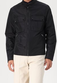 Černá bunda na zip s vysokým límcem, vybavená dvěma náprsními kapsami a bočními kapsami na zip. Vyrobena z hladké tkaniny s matným povrchem.